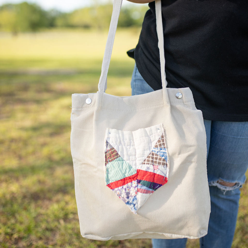 A person holding a beige tote bag with a unique quilt pattern on the front pocket, standing in an outdoor setting.