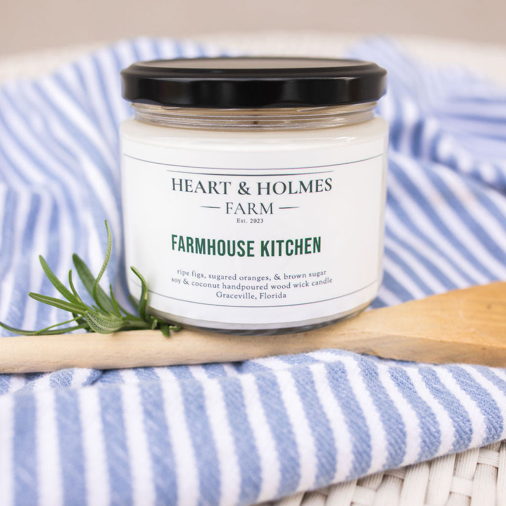 A jar of 'Farmhouse Kitchen' wax tart by Heart & Holmes Farm, placed on a blue striped cloth beside a wooden spoon.