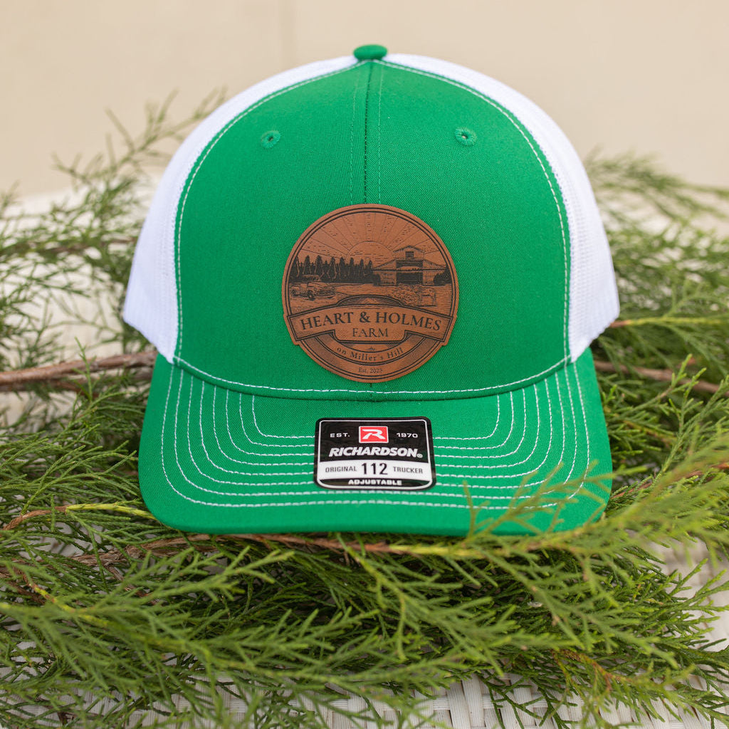 A green and white leather patch baseball cap with an engraved logo on the patch, displayed on a wicker surface with pine needles around it.