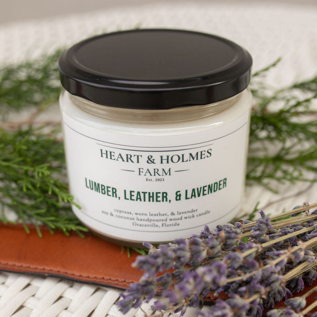 A candle with a label reading 'Heart & Holmes Farm Lumber, Leather, & Lavender' placed on a wicker table accompanied by a small bundle of dried lavender and a leather-bound notebook.
