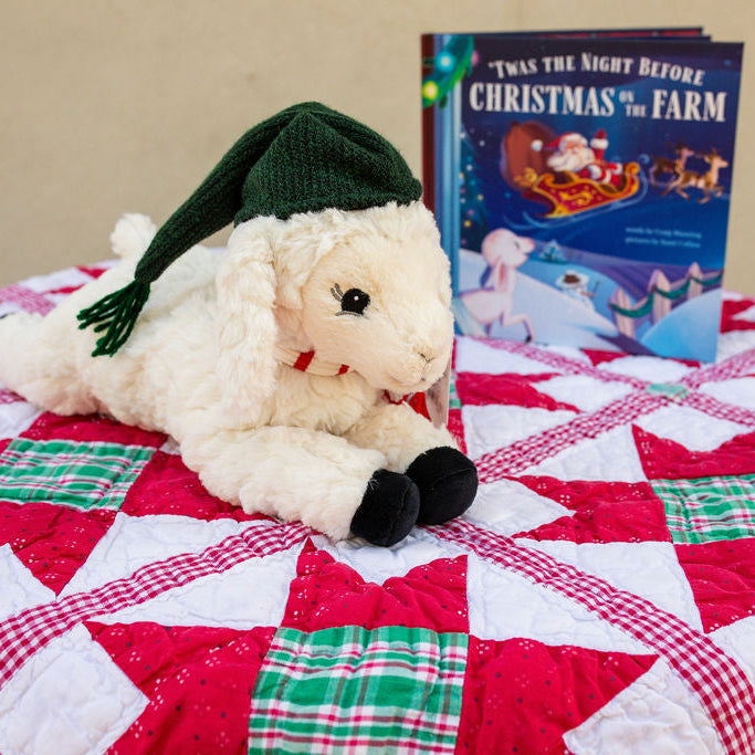 Stuffed sheep with a green hat on a quilt next to a Christmas-themed book